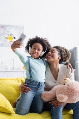 Afro-Amerikan kulaklıklı kız ve oyuncak ayılı kadın dil çıkarırken selfie çekiyorlar.