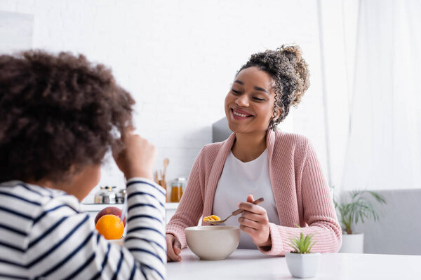 улыбающаяся африканская американка, держащая ложку с кукурузными хлопьями, глядя на дочь на размытом переднем плане