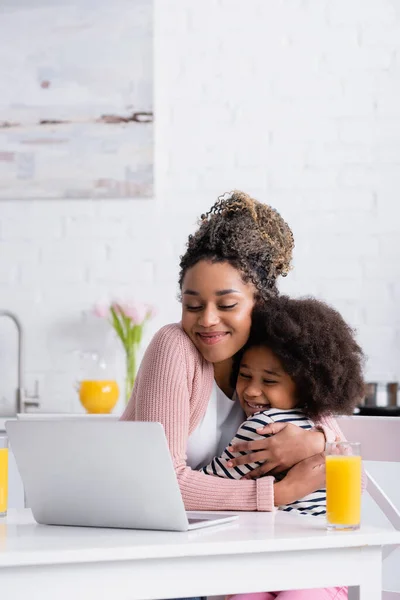 happy african american mom embracing daughter while watching film on laptop during breakfast