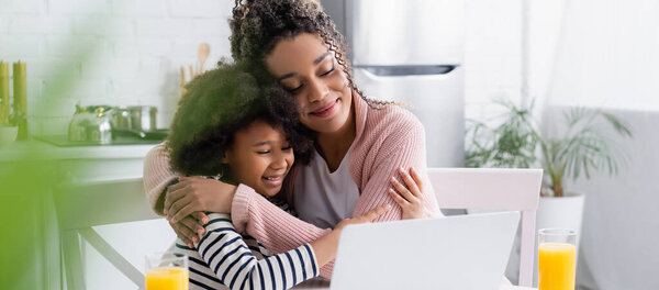 happy african american mom and daughter embracing while watching film on laptop, blurred foreground, banner