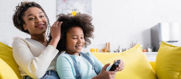 テレビやバナーを見ながらリモコンを持っている娘の髪に触れる幸せなアフリカ系アメリカ人女性 — ストック写真