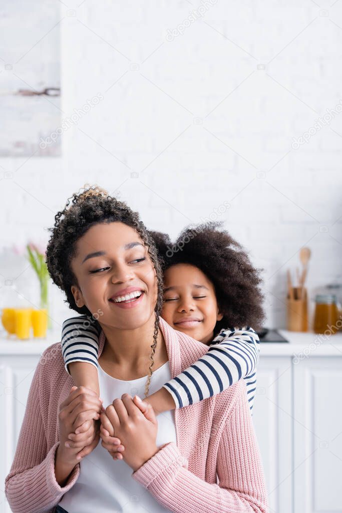 happy african american girl with closed eyes holding hands of cheerful mother in kitchen