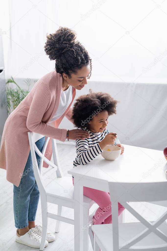 smiling african american woman touching shoulders of daughter having breakfast in kitchen