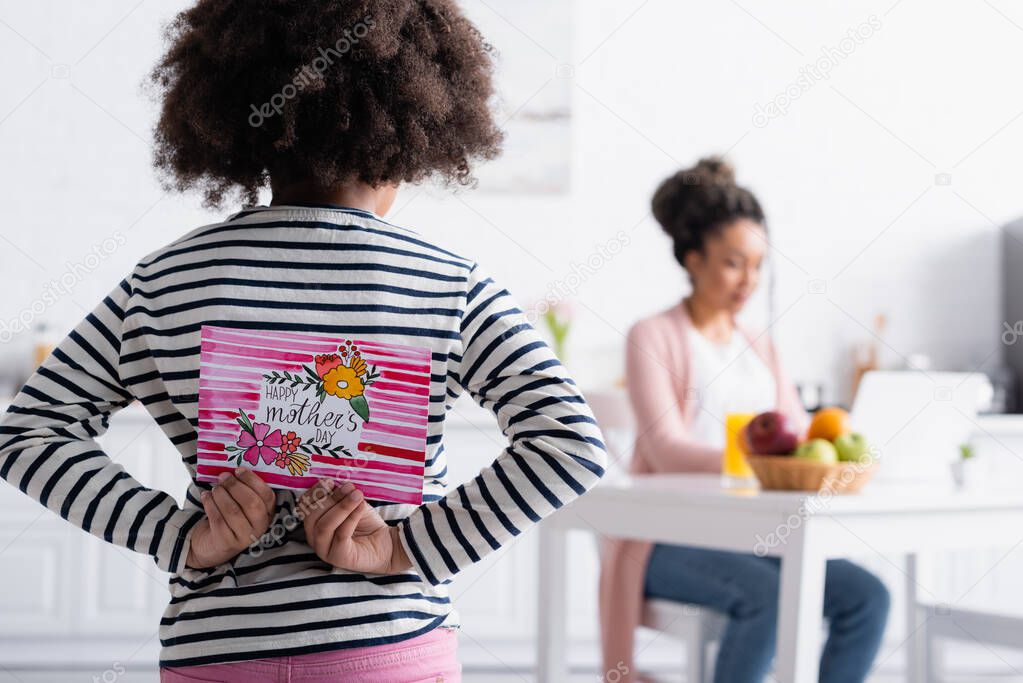 back view of african american child holding happy mothers day card near mom working in kitchen on blurred background