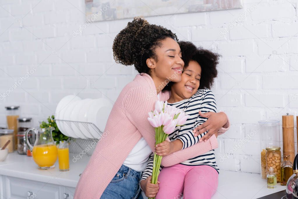 pleased african american woman embracing daughter sitting on kitchen counter with tulips