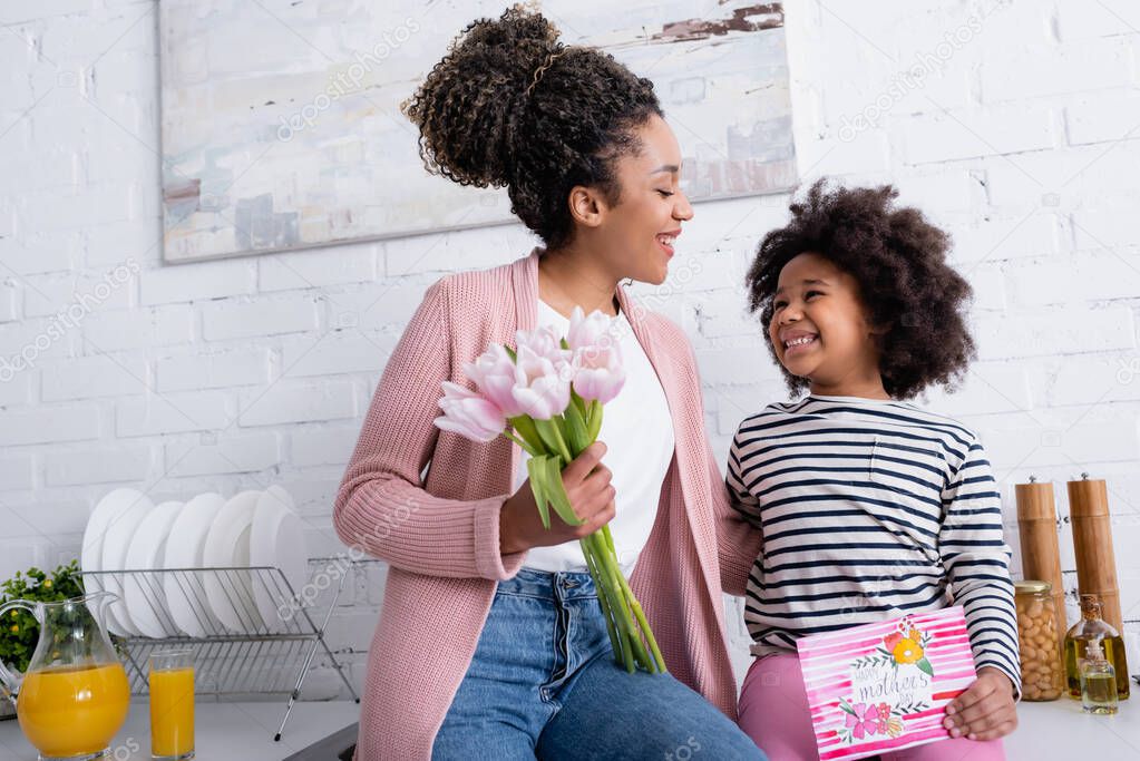 happy african american woman holding tulips near cheerful daughter with happy mothers day card