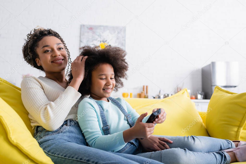 happy african american woman touching hair of daughter holding remote controller while watching tv