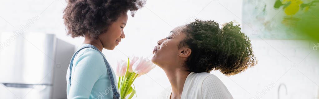 side view of african american woman blowing air kiss to smiling daughter with fresh tulips, banner