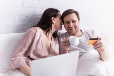 Woman whispering to smiling husband with credit card and coffee near laptop on blurred foreground on bed  clipart