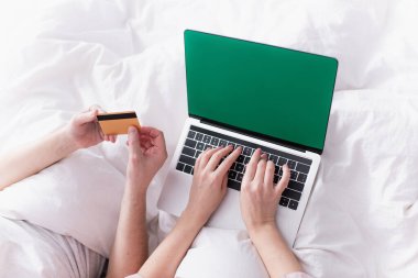 Yatağında kredi kartı olan, kocasının yanında krom anahtarlı dizüstü bilgisayar kullanan kadın görüntüsü. 
