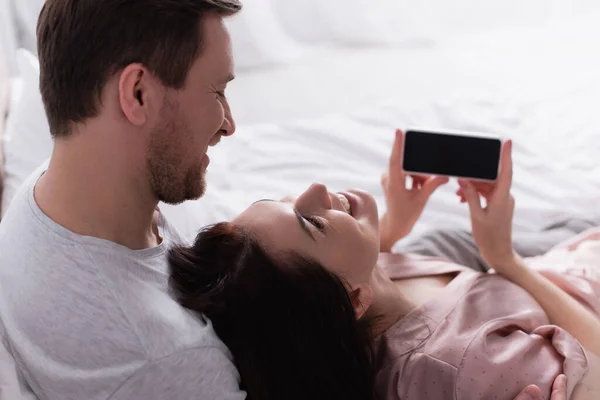 笑顔の女性はベッドの上に空白の画面でスマートフォンを保持しながら夫を見て — ストック写真