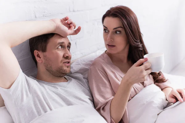 拿着咖啡和智能手机的女人躺在床上看着丈夫 — 图库照片