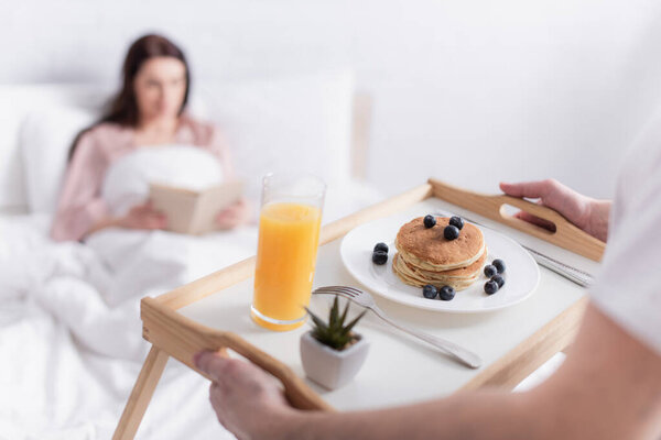 Man holding breakfast on tray near wife on blurred background in bedroom 