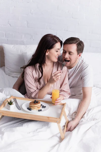 Glimlachende Vrouw Voedt Man Buurt Van Pannenkoeken Sinaasappelsap Bed — Stockfoto