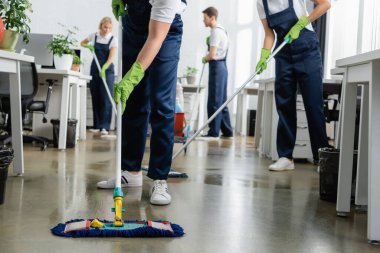 Cleaner cleaning floor with mop near colleagues working on blurred background in office  clipart