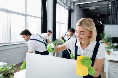 Cleaner smiling while cleaning computer monitor with detergent in office  clipart