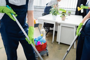 Cropped view of cleaners in overalls holding mop and vacuum cleaner near cart with detergents in office  clipart