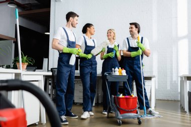 Smiling multiethnic cleaners talking near cart with detergents in office  clipart