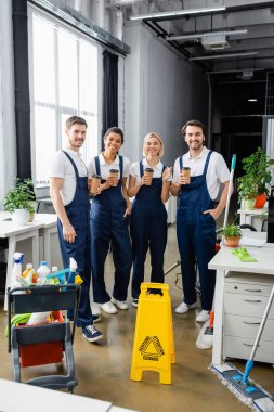 Cheerful multiethnic cleaners holding coffee to go near detergents in office  clipart