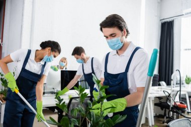 Tıbbi maskeli bir temizlikçi, bulanık arka planda bitki ve çok ırklı meslektaşların yanında deterjan tutuyor. 