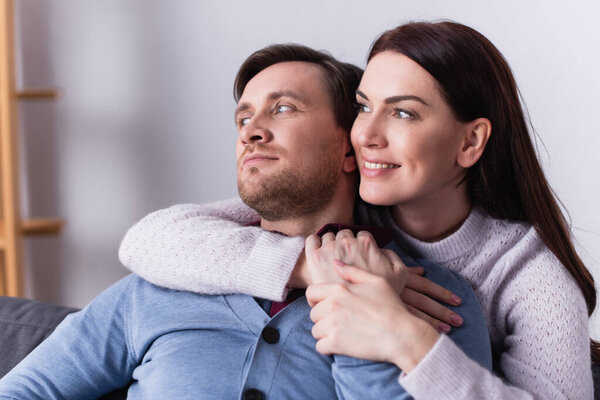 Adult woman hugging husband and looking away at home 