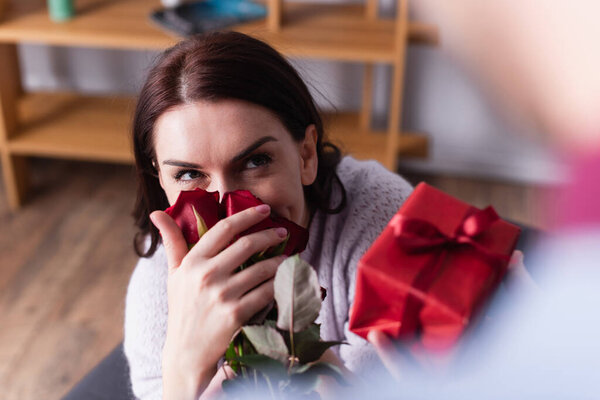 Brunette woman smelling flowers near husband with gift on blurred foreground 