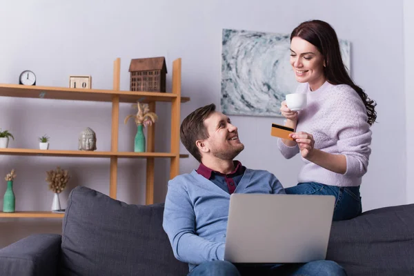 Mujer Con Taza Tarjeta Crédito Mirando Marido Con Portátil Sofá — Foto de Stock