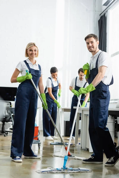 Limpiadores Sonrientes Lavando Piso Cerca Colegas Multiétnicos Con Fondo Borroso — Foto de Stock