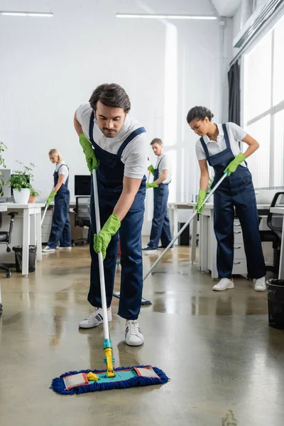 Cleaner in overalls washing floor near multiethnic colleagues in office