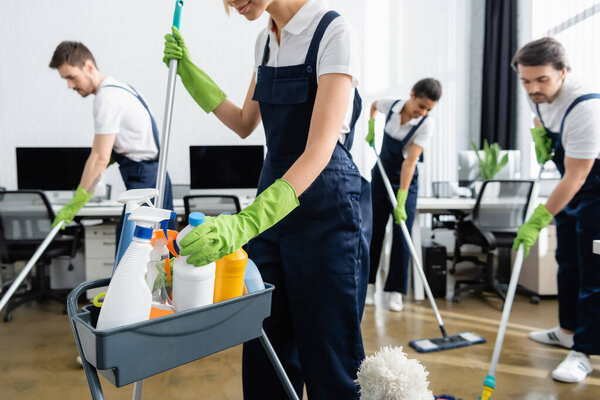 Cleaner in uniform holding mop and taking detergent near colleagues in office 