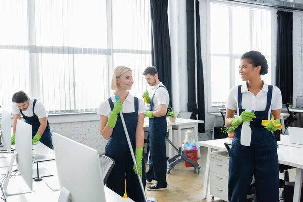 Interracial colleagues smiling at each other while holding cleaning supplies in office 