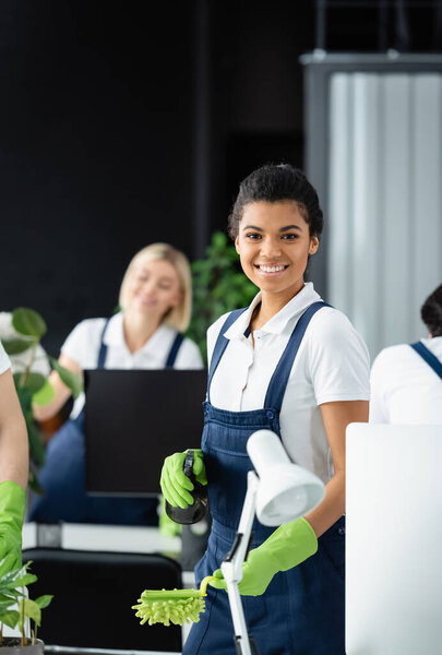 Smiling african american cleaner with dust brush and detergent looking at camera in office 