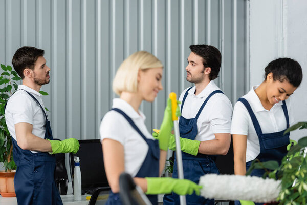 Workers of cleaning service talking near smiling multiethnic colleagues on blurred foreground in office 