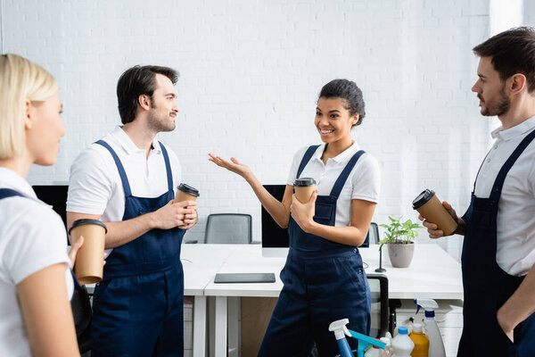 African american cleaner holding paper cup while talking with colleagues in office 