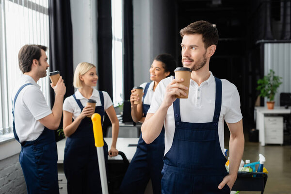 Cleaner in overalls holding coffee to go near interracial colleagues talking on blurred background 
