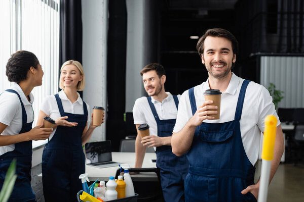 Positive cleaner holding paper cup and looking at camera near interracial colleagues and detergents in office 