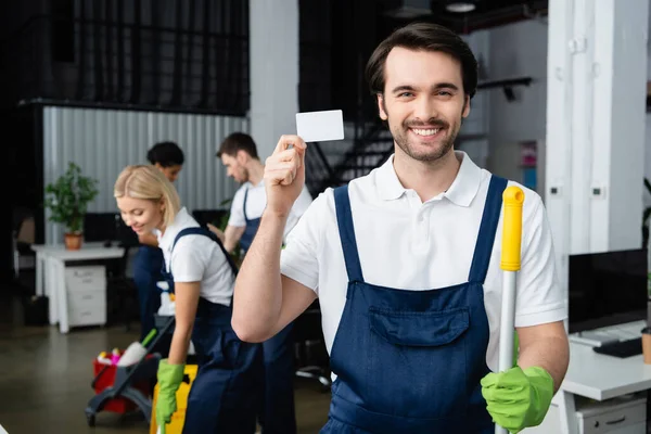 Positive Cleaner Holding Empty Card Mop While Colleagues Working Office — Stock Photo, Image