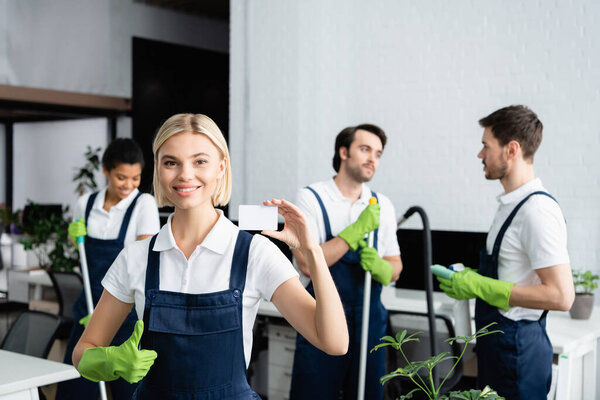 Positive cleaner with empty card showing like near multiethnic colleagues in office 