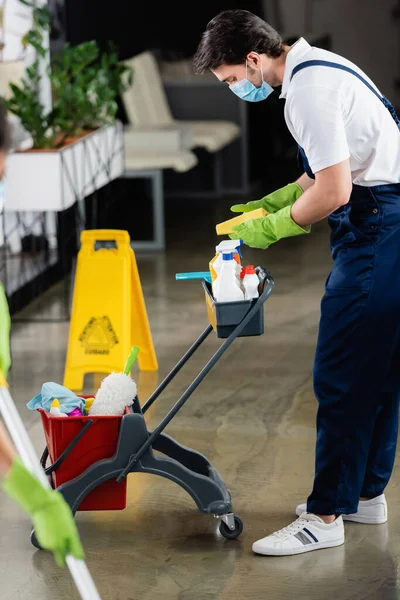 Worker of cleaning service in medical mask holding rag near cart with detergents in office