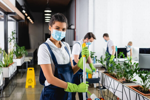 African american cleaner in medical mask holding mop while colleagues working on blurred background in office 