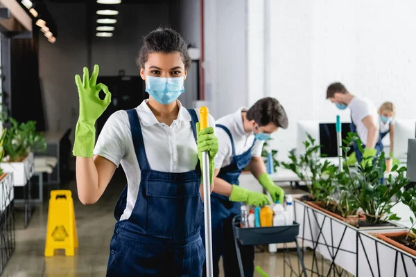 African american worker of cleaning company in medical mask holding mop and showing ok gesture in office