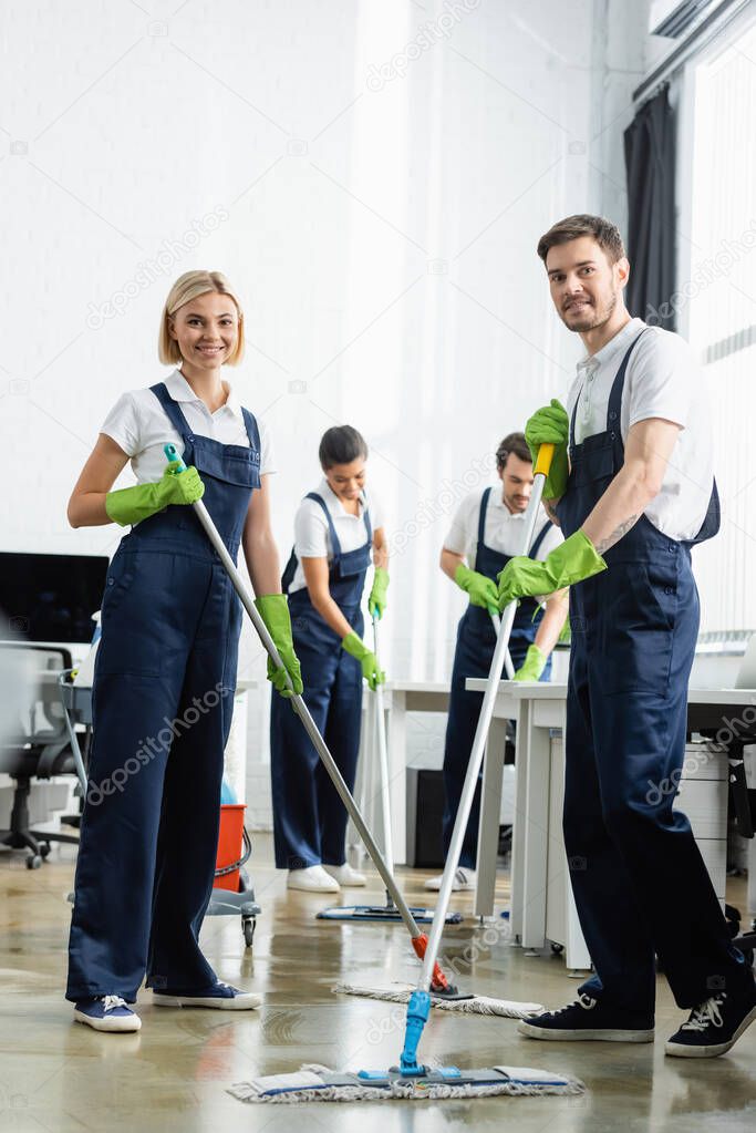 Smiling cleaners washing floor near multiethnic colleagues on blurred background in office 