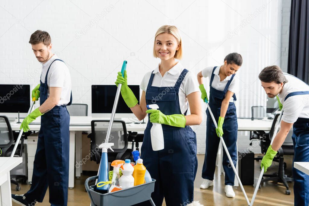 Smiling cleaner holding detergent and mop near multiethnic colleagues in office 