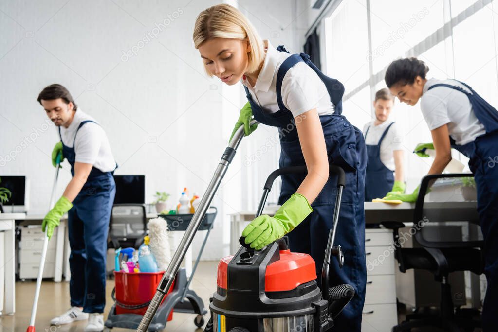Cleaner in overalls using vacuum cleaner near multiethnic colleagues in office 