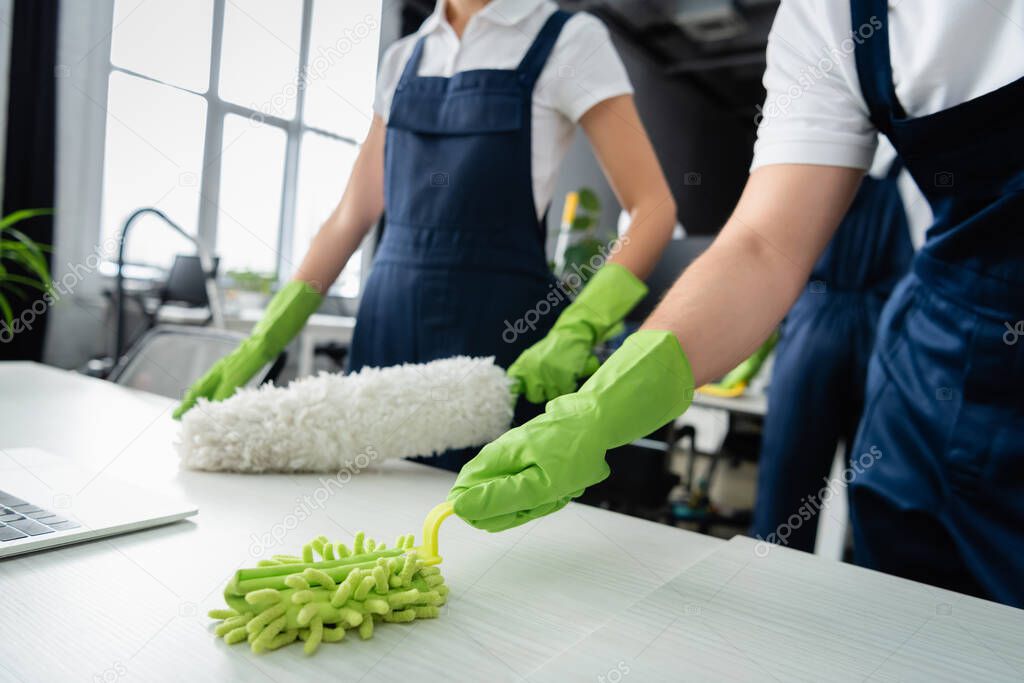 Cropped view of cleaner cleaning table near colleague with dust brush in office 