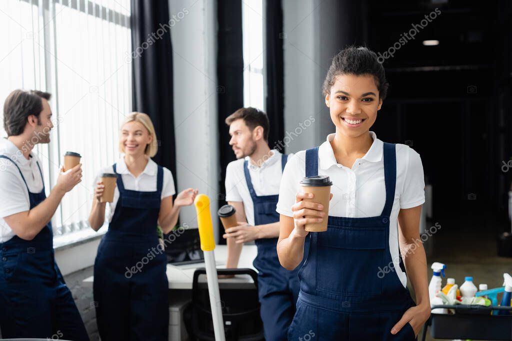 African american cleaner holding paper cup near colleagues on blurred background in office 