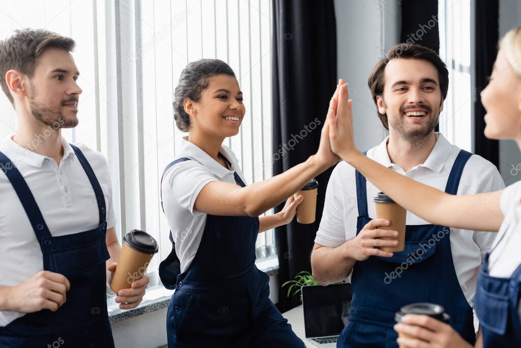Interracial cleaners giving high five near colleagues with coffee to go in office 