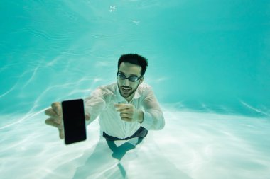 Gülümseyen Müslüman işadamı su altında bulanık akıllı telefonu işaret ediyor. 