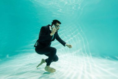 Muslim businessman in suit talking on smartphone underwater in pool  clipart