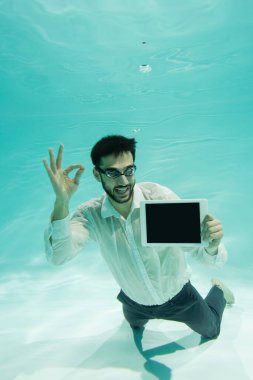 Gülümseyen Müslüman işadamı iyi niyet gösteriyor ve su altında dijital tablet tutuyor. 
